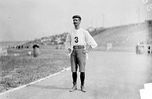 Black and white photograph of man and hat with mustache wearing a white shirt with the number three on it and trousers on a smooth path adjacent to cut grass near empty stands