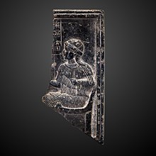Bas-relief en pierre noire représentant une femme assise, vêtue d'une tunique rayée et d'un bonnet.