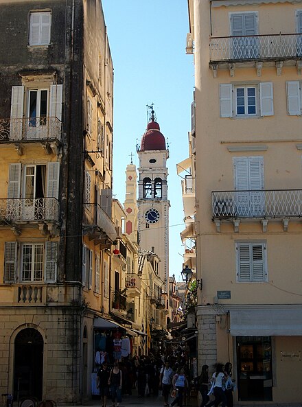 The bell tower of the Saint Spyridon Church can be seen in the background among the busy kantounia of the city centre. On top of the stores are apartments with balconies. It is from these type of balconies that Corfiots throw botides, clay pots, to celebrate the Resurrection during Easter festivities.