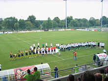 Zwei Fußballmannschaften, eine gelb und schwarz, die andere hellgrün, reihen sich auf einem Sportplatz auf.  Im Hintergrund: Reklametafeln und Bäume