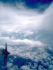 Una fotografía del ojo de un huracán.  Uno de los motores de un avión cazador de huracanes se puede ver en la parte inferior derecha de la imagen.  Las nubes dispersas en la parte inferior dan paso al agua.  En el medio, se ve claramente la pared del ojo de la tormenta y cerca de la parte superior, hay tenues nubes cirros.  Una parte de la parte superior de la imagen muestra el cielo.