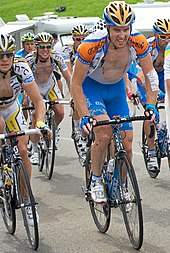A group of road racing cyclists in various jerseys, with expressions of pain and fatigue on their faces.