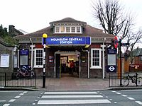 A brown-bricked building with a rectangular, blue sign reading "HOUNSLOW CENTRAL STATION" in white letters and a light post in front all under a white sky