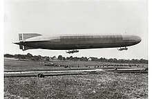 Schwarz-Weiß-Foto eines Luftschiffs in Bodennähe. Darunter sind zahlreiche Personen auf einer Wiese erkennbar.