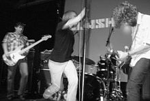 Ein Foto einer Rockband, Mudhoney, bei einer Live-Show.  Das Foto ist durch die Bewegung auf der Bühne unscharf.  Von links nach rechts sind der E-Bassist, Sänger und Gitarrist.