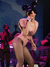 Eine Frau, die ein hoch tailliertes rosa funkelndes Outfit mit großen Schulterpolstern trägt, die auf einer Bühne auftreten