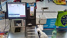 A self-service checkout at a Tesco store in Poland; a barcode scanner is in the glass below the display screen; below this is a flat metal plate on which produce may be weighed; a bank card PIN pad is to the right of the display screen; and to the right is the bagging area Self-service checkout in Tesco in Poland.jpg