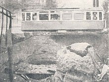 A tram car occupies the top half of the picture. The bottom half shows a several-metre-deep ditch containing soil, boulders, and torn cables or piping.