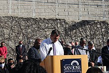San Jose Mayor Sam Liccardo stands on a stage with his head down with everyone around him in the audience doing the same.