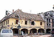 Zwei zerschmetterte Arkadenhäuser, eines alt und gelb gestrichen, das andere moderner und aus Backstein und Beton, mit zerstörten Dächern und vielen Einschusslöchern.  Davor ist eine beschädigte Ampel und ein Auto.