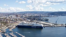 Foto einer direkt am Meer gelegen Stadt, im Vordergrund ein großes Schiff. Direkt an der Küste befindet sich vor allem Verkehrsinfrastruktur