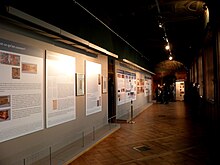 Longue galerie sombre éclairée de fenêtres par la droite et présentant de grands panneaux sur le mur de gauche