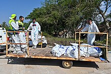 Four men in white garb, standing among body bags, on a moving platform.