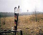 View across a landscape with a leaning sign reading "Halt Hier Zonengrenze Bundesgrenzschutz" in the foreground, a red/black/yellow striped square-shaped pole just behind, and a metal fence and watchtower visible across a strip of open ground in the background.
