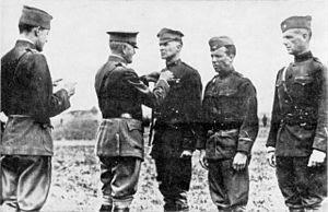 Three men in uniform are standing side by side. The one on the left is wearing a peaked "crush cap" and standing smartly at attention, while the two on the right wear garrison caps and are slouching. A man in a peaked cap and Sam Browne belt is pinning something on the chest of the first man. Behind him stands another man in a garrison cap who is reading a document in his hands.