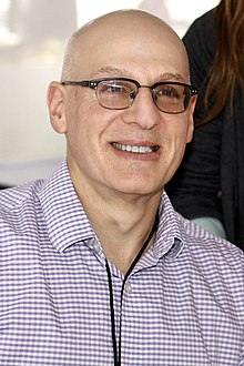 Black and white photograph of Gordon Korman, author of young adult fiction, speaking at the National Book Festival in September 2011. Photograph depicts Korman in profile view, facing left and speaking into a microphone. His right hand is raised to approximately shoulder height, palm facing the audience, with fingers slightly closed as if grasping an invisible ball.