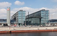 Frontale Farbfotografie auf das Glasgebäude. Direkt an der Uferpromenade stehen zwei sechs geschossige Teilgebäuden mit einer kleineren Glasfront in der Mitte, die als Aufschrift „DB Berlin Hauptbahnhof“ hat. Alle drei Gebäude sind von einer großen Glasröhre durchzogen. An der linken Seite steht ein grauer Turm mit dem DB Logo.