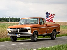 Un pick-up Ford F-250 de couleur orange avec une plaque d'immatriculation allemande. Le véhicule est en train de rouler avec un homme au volant, un drapeau américain est fixé sur le bord de son plateau et vole au vent.