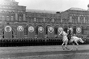 Photo noir et blanc avec au premier plan un homme galopant sur un cheval blanc, au second des soldats au garde-à-vous devant un grand bâtiment décoré de bannières.