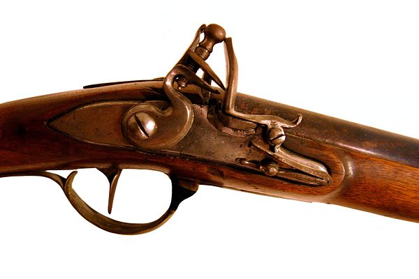 This flintlock mechanism is distinct from the metal barrel extending to the right, and the surrounding wooden stock encloses and obscures the trigger 