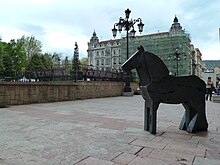 Фотография стилизованной геометрической статуи пони на площади со старым строящимся зданием на заднем плане.