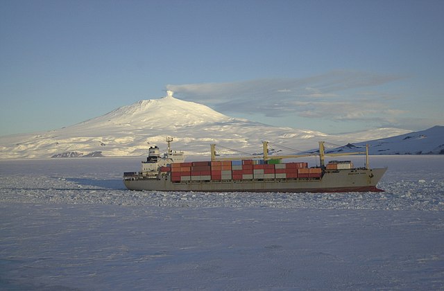 Photographie d'un bateau traversant la banquise, et en arrière un volcan fumant, couvert de neige.