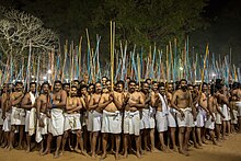 Uma tropa de homens, na frente, vestidos com uma tanga branca, descalços, brandem grandes gravetos.