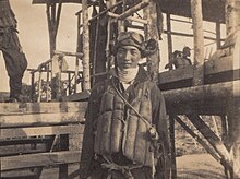 Photo noir et blanc d'un homme en tenue de pilote devant une grande structure en bois.