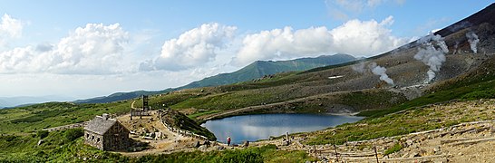 Eine Panorama-Fotografie zeigt eine imposante Landschaft im Gebirge. In der Mitte ist ein kleiner See und links davon ein Steinhaus zu sehen. Der Himmel ist strahlend blau.