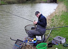 Man seated at the side of the water surrounded by fishing rods and tackle.