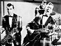 A black-and-white photograph of three men playing musical instruments (a saxophone, a guitar, and a drum kit), the drummer in the background