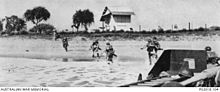 Des soldats débarquent sur une plage, se précipitant vers le bâtiment en bordure de plage.
