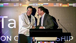 Two men on a stage smiling and shaking hands, the one at the podium is wearing a business suit and the other is wearing a white hoodie