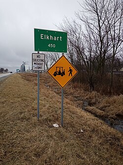 Großes grünes Schild im Vordergrund mit der Aufschrift "Elkhart, 450", montiert auf zwei Metallpfosten, mit einem kleineren weißen Schild mit der Aufschrift "Keine Werbung ohne Erlaubnis" und einem gelben rautenförmigen Schild mit der Zeichnung eines Gabelstaplers und einer gehenden Person;  Getreideheber im Hintergrund