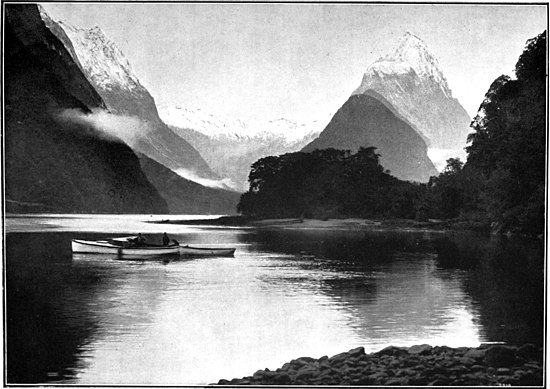 Small boats on a very still lake-like area, surrounded by snow-capped mountain peaks with steep slopes that come down to the water's edge, with bush and trees around the water's edge on the right