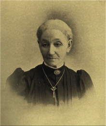 B&W portrait photo of a woman with her white hair in an up-do, wearing a dark blouse, with puffy sleeves and white fringe at the collar, as well as brooch at the neckline and a necklace.