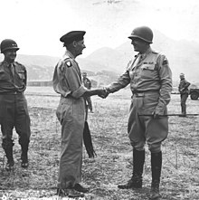 Patton souriant tenant une cravache et portant de hautes bottes et son casque serre la main de Montgomery qui porte un béret.