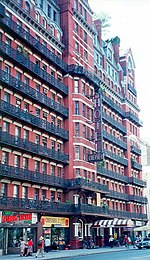 Einige Fußgänger laufen am Chelsea Hotel vorbei, einem mehrstöckigen, rot-weißen Gebäude mit einer großen Anzahl von Zimmern inklusive Balkonen pro Etage. Erneut steht der Gebäudename auf einer vertikalen Tafel, zudem an einer Markise im Erdgeschoss.