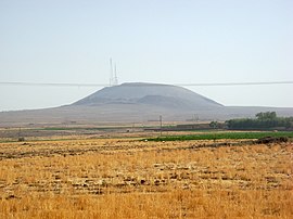 Ein schwarzer Berg mit einem Telekommunikationsturm darüber, von weitem gesehen und durch staubigen Nebel verzerrt. Sie stehen auf einem Feld, das von trockenen Gräsern bewachsen ist, von denen einige grün sind.