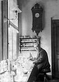 An old, bespectacled man wearing a suit and sitting at a bench by a large window. The bench is covered with small bottles and test tubes. On the wall behind him is a large old-fashioned clock below which are four small enclosed shelves on which sit many neatly labelled bottles.