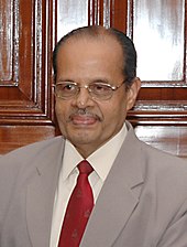 Photograph of an old man wearing a suit with red-coloured tied and glasses.