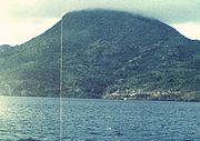 A photograph depicting a blue sky at the top, a grey mountain range in the middle, white clouds in front of the mountain range, and a rocky terrain at the bottom.