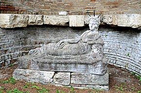 Reclining Attis with radiate crown, holding a shepherd's crook (damaged) in his left hand and in his right pomegranates, pine cones, and wheat: his partial nudity shows that he has undergone complete castration, and the bearded head on which he leans is most likely the river god Sangarius or Gallus (from Ostia, 2nd century AD) Statue of a reclining Attis at the Shrine of Attis 1.jpg