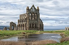 Foto av en stor ødelagt, takløs kirke med utsikt over en dam i forgrunnen og havet i horisonten