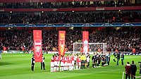 A line of people on a field, behind them are three red banners. In the background is a stand with three tiers