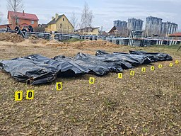 [Foto: Zehn schwarze, durchnummerierte Leichensäcke liegen auf dem Boden; im Hintergrund links Einfamilienhäuser und rechts Hochhäuser; Szene nach der Entdeckung des Massakers von Butscha im April 2022]