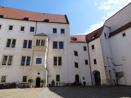 The inner courtyard of Colditz castle which was used as the prison yard when the castle was the POW camp Oflag IV-C during World War II. The door flanked by bushes was the entrance to the "Prominente" quarters. Note the cutout depiction of Lieutenant Bouley to the lower left hand side of the photograph.