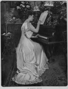 A white woman seated at a piano, one hand on the keys and one hand writing on a musical score; she is wearing a long light-colored gown; there are bouquets in the background