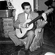 Photographie d’un homme en costume, assis dans un fauteuil à droite d’une cheminée, jouant de la guitare.