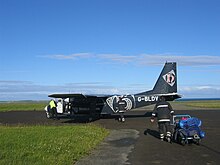 Een klein donkerblauw twinpropvliegtuig zit op asfalt omringd door gras onder een blauwe lucht.  Op de voorgrond trekt een persoon in een uniform dat qua kleur lijkt op het vliegtuig een volle bagagewagen naar zich toe.
